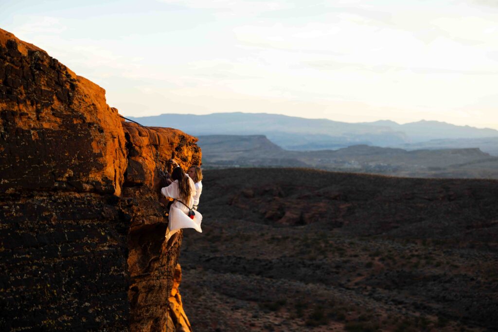 Couple gearing up in wedding attire with climbing harnesses at the base of a massive rock wall in Kanab, Utah.