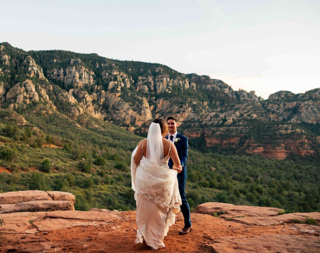 Newlyweds holding hands, walking along a scenic Sedona trail at sunset
